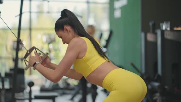 Eine junge hispanische Frau im gelben Trainingsanzug führt eine Übung in einem Crossover durch und zieht ein Stahlseil von oben, um ihren Rücken und ihre Schultern zu trainieren. Eine Frau trainiert Rücken und Schultern in einer Turnhalle - Filmmaterial, Video