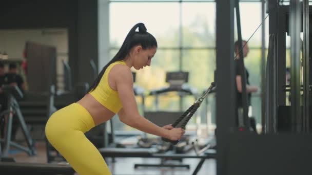 Eine junge hispanische Frau im gelben Trainingsanzug führt eine Übung im Crossover durch, bei der sie ein Seil von oben zieht, um Rücken und Schultern zu trainieren. Brünette Frau trainiert Rücken und Schultern in der Turnhalle - Filmmaterial, Video