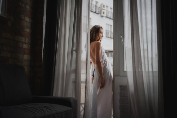 Bela sensual jovem com cabelos longos em uma camisa branca em pé junto à janela com cortinas - Foto, Imagem