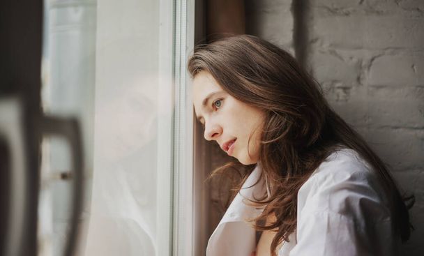 Portret van een jonge mooie sexy vrouw in een wit shirt zittend bij het raam in een appartement - Foto, afbeelding
