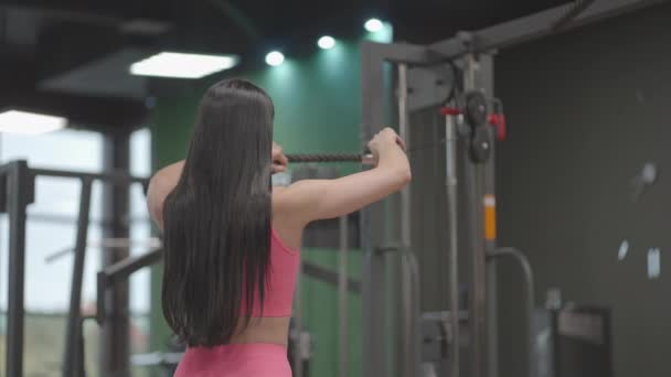 A brunette woman in a pink suit pulls a rope in a crossover trainer to her chest. Back workout in a trainer. Professional woman instructor - Footage, Video