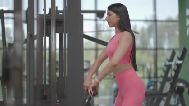 Een Latijns-Amerikaanse brunette in een roze pak trekt een touw in een crossover met haar handen om haar schouders te trainen. Schoudertraining in een trainer. Professionele vrouwelijke instructeur - Video