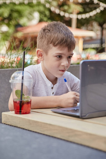 Kleiner Schüler macht Hausaufgaben im Freien mit Laptop, schreibt Notizen ins Textbuch - Foto, Bild
