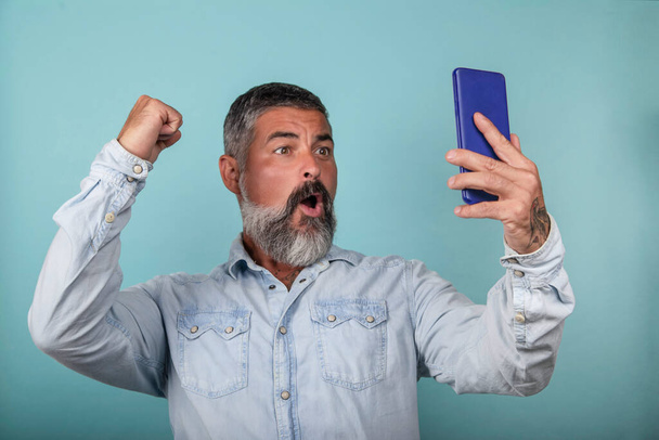 Zdjęcie triumfującego szczęśliwca, trzymającego telefon komórkowy i wykonującego gest zwycięzcy stojącego nad jasnoniebieskim tłem - Zdjęcie, obraz