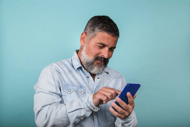 Hombre barbudo sonriente y bien parecido usando un teléfono inteligente para contactar a su familia y amigos aislados sobre un fondo azul - Foto, Imagen