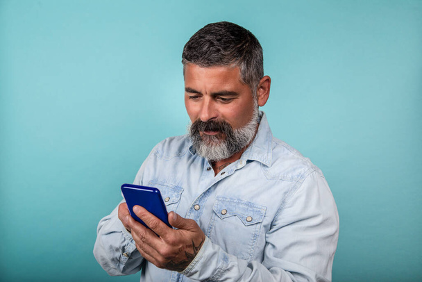 Foto eines fröhlichen bärtigen Mannes, der sein Smartphone auf blauem Hintergrund benutzt - Foto, Bild