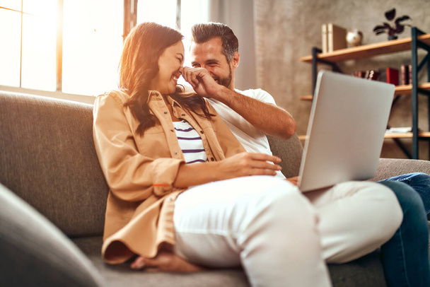 Pareja joven enamorada están sentados en el sofá con un ordenador portátil en casa. Una pareja casada se está relajando y divirtiendo en la sala de estar. Compras en línea, trabajo desde casa. - Foto, imagen