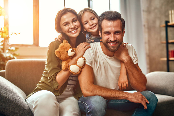 Happy dad and mom with their cute daughter and teddy bear hug and have fun sitting on the sofa in the living room at home. - Photo, Image
