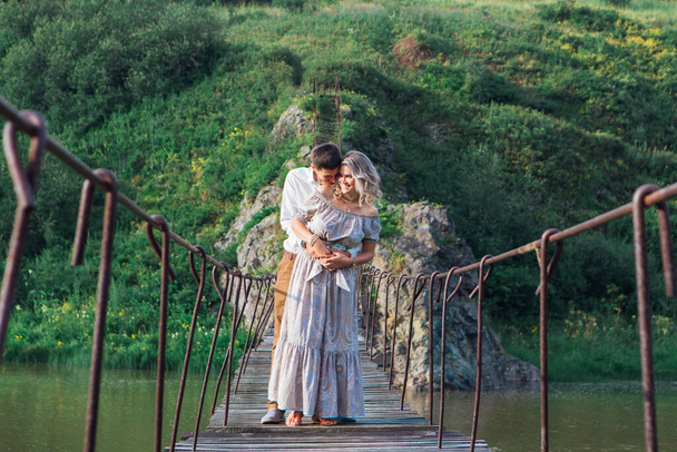 Beautiful young romantic couple standing and hugging on the suspension bridge above the river - Foto, afbeelding