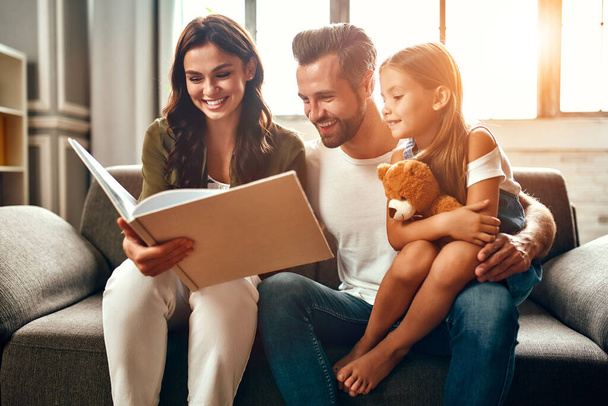 Happy dad and mom with their cute daughter and teddy bear are reading a book while sitting on the sofa in the living room at home. - Foto, imagen