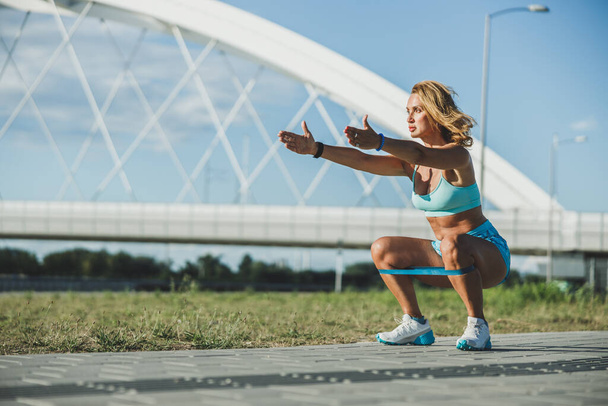Adult woman doing squats with resistance band on her legs while working out near the river. - Foto, Imagem