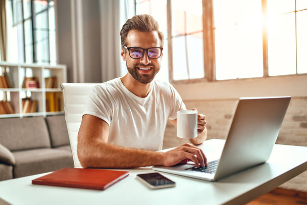Homme d'affaires dans des lunettes avec une tasse de café fonctionne à un ordinateur portable. Freelance, travail à domicile. - Photo, image