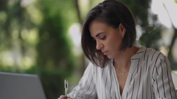 Business Woman Brunetka Arabska Latynoska Pochodzenie etniczne Działa zdalnie siedząc w letniej kawiarni w słoneczny dzień z laptopem i zapisuje długopisem i notatnikiem - Materiał filmowy, wideo