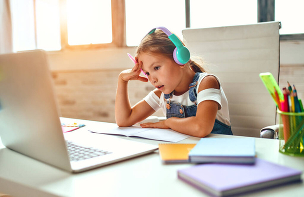 オンライントレーニング。ヘッドフォンの子供の女の子はノートパソコンでレッスンを聞く。パンデミックや隔離された家庭での学校. - 写真・画像