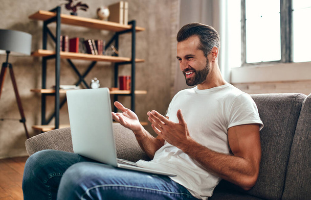 Un homme est assis sur le canapé avec un ordinateur portable sur les genoux. Homme d'affaires sur vidéoconférence fonctionne en ligne à partir de la maison. - Photo, image