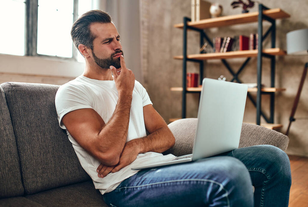 Мужчина задумчиво сидит на диване с ноутбуком на коленях. Бизнесмен работает онлайн из дома. Фриланс, онлайн-шопинг. - Фото, изображение