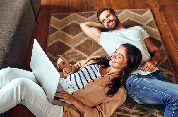 Fiatal, álmodozó pár fekszik otthon a szőnyegen. Egy házaspár pihen a nappaliban. Egy nő laptoppal a földön fekszik egy férfin. Otthonról dolgozom. Online vásárlás. - Fotó, kép