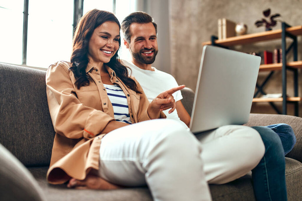 A szerelmes fiatal pár otthon ül a kanapén egy laptoppal. Egy házaspár pihen a nappaliban. Online vásárlás, otthoni munka. - Fotó, kép
