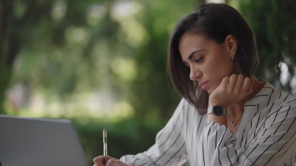 Business Nainen Brunette arabia latinalaisamerikkalainen etnisyys toimii etänä istuessaan kesäkahvilassa aurinkoisena päivänä kannettavan tietokoneen kanssa ja kirjoittaa kynällä ja muistikirjalla - Materiaali, video