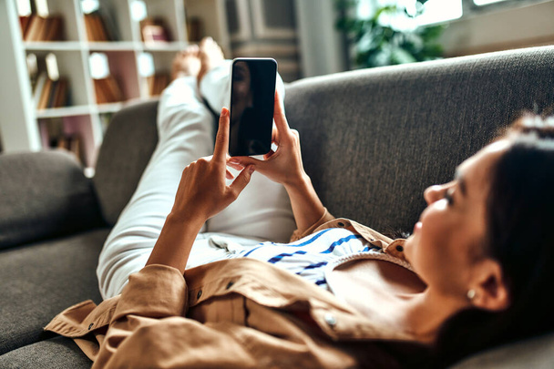 Egy nő fekszik a kanapén egy telefonnal a nappaliban. Kommunikáció, online vásárlás. Egy okostelefonos nő kommunikál, pihen és szórakozik. Okostelefon üres képernyő. - Fotó, kép
