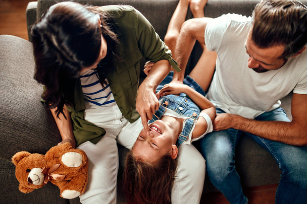 Happy dad and mom with their cute daughter and teddy bear hug, play and have fun sitting on the sofa in the living room at home. - Photo, Image