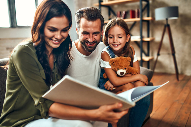 Щасливий тато і мама зі своєю милою дочкою і плюшевим ведмедем читають книгу, сидячи на дивані у вітальні вдома
. - Фото, зображення