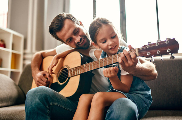 幸せなお父さんは、自宅のリビングルームのソファに座っている間にギターを弾くために彼のかわいい娘を教えています。幸せな父の日. - 写真・画像