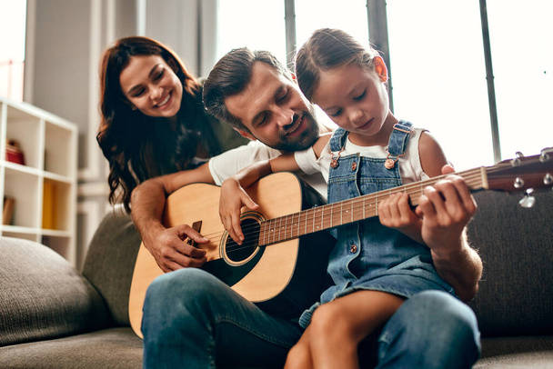 Glückliche Väter und Mütter bringen ihrer süßen kleinen Tochter das Gitarrespielen bei, während sie zu Hause auf dem Sofa im Wohnzimmer sitzen. Die Familie verbringt Zeit miteinander. - Foto, Bild