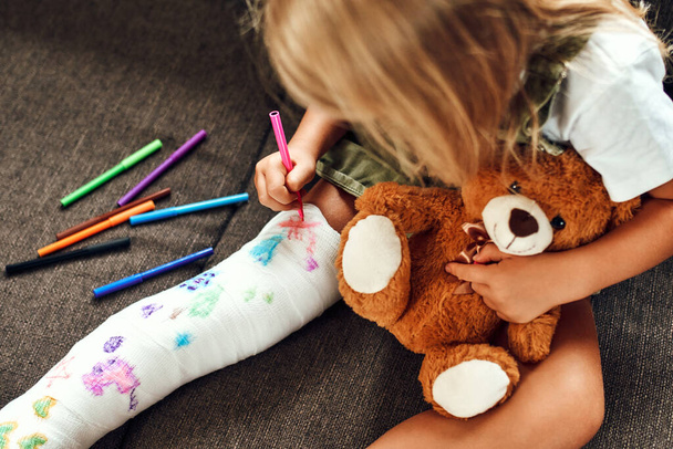 Menina com uma perna partida no sofá. A criança desenha com canetas de hidrográfica em uma atadura de bandaid. - Foto, Imagem