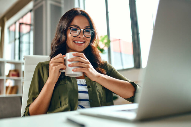 Business woman works at a laptop while sitting at a table at home and drinks coffee. Freelance, work from home. - Photo, Image