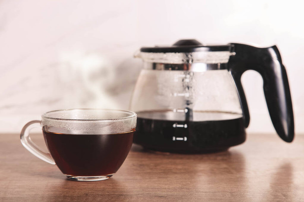 Горячий кофе в стеклянной чашке и кувшин кофеварки на деревянной кухонной столешнице.  - Фото, изображение