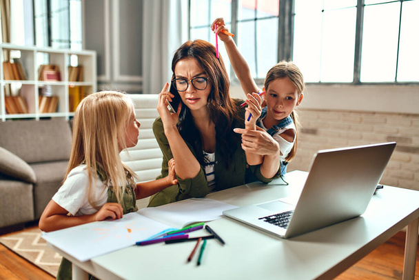 Een zakenvrouw en een moeder proberen aan een laptop te werken als haar dochtertjes spelen, rotzooien en zich met haar bemoeien. Freelance, thuiswerk. - Foto, afbeelding