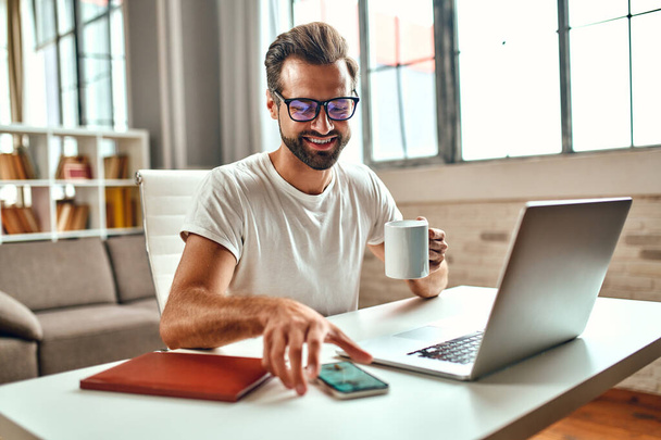 Businessman in glasses with a mug of coffee works at a laptop. Freelance, work from home. - Foto, afbeelding