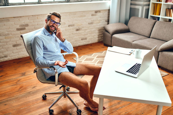 Een man in een hemd en korte broek zit aan een tafel en praat over een videogesprek, een online zakelijke conferentie, een vergadering. Freelance, thuiswerk. - Foto, afbeelding