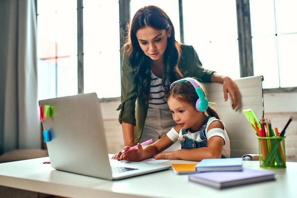 Online training. Mam helpt haar dochter met lessen. Schoolmeisje in koptelefoon luistert naar een les op een laptop. School thuis in een pandemie en quarantaine. - Foto, afbeelding