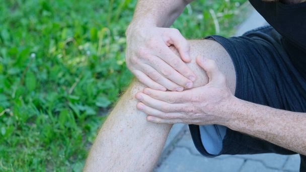 Мужчина бегун трогает ногу от боли из-за растяжения лодыжки. Неузнаваемый спортсмен сидит на земле и страдает от боли в колене - Фото, изображение