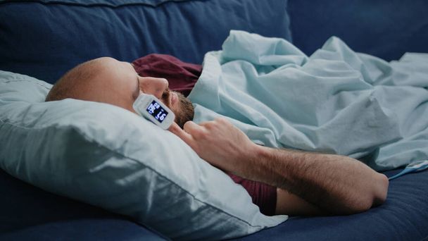 Крупним планом хворий чоловік спить на дивані з оксиметром на пальці
 - Фото, зображення