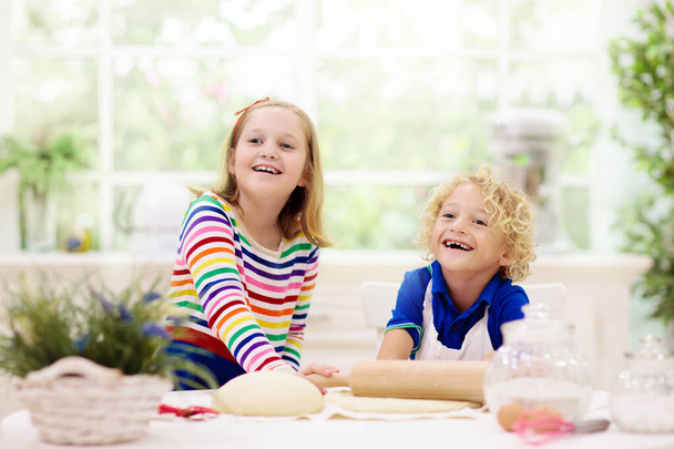 Děti pečou koláč v bílé slunné kuchyni. Děti vaří večeři. Chlapec a dívka valí těsto na velké okno. Zdravá výživa pro dítě. Bratr a sestra si hrají doma. - Fotografie, Obrázek