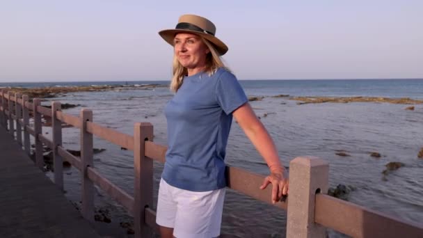 Smiling middle aged woman wearing blue t-shirt and straw hat and standing at sunset on the wooden bridge with hands on it. T-shirt mockup - Footage, Video