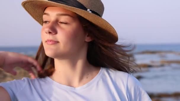 Tini lány szalmakalapban áll a tenger mellett naplementekor, félrenéz és mosolyog. Fejlövés, közelről. Nyári utazási koncepció.  - Felvétel, videó