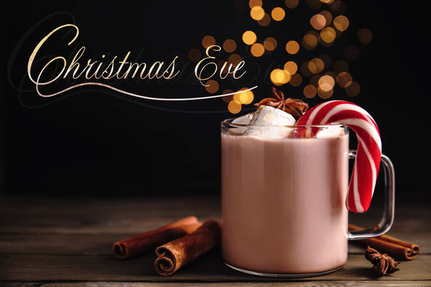 Christmas Eve, postcard design. Glass cup of tasty cocoa with marshmallows and candy cane on wooden table against blurred festive lights - Photo, image