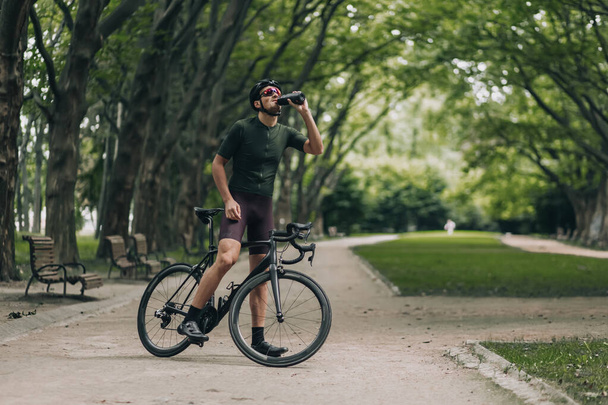 Καυκάσιος μυώδης άνδρας πίνει φρέσκο νερό από το αθλητικό μπουκάλι ενώ ξεκουράζεται μετά το πρωινό ποδήλατο στο καλοκαιρινό πάρκο. Έννοια του αθλητισμού, αναψυχή και δροσιστικό. - Φωτογραφία, εικόνα