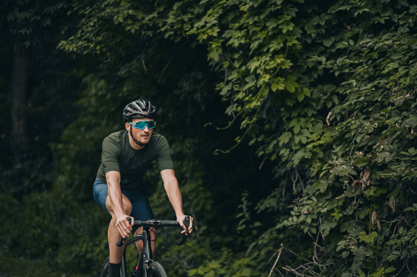 Kasklı ve aynalı gözlüklü sporcu temiz havada bisiklet sürmeye çalışıyor. Yeşil şehir parkında siyah bisiklet süren Kafkas sporcusu. - Fotoğraf, Görsel
