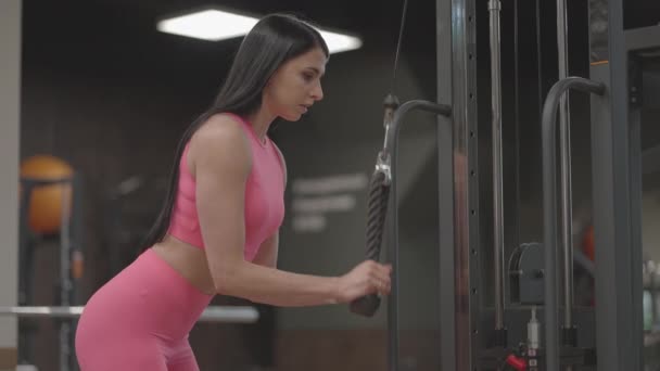 Eine brünette Frau im rosafarbenen Anzug zieht ein Seil in einem Crossover-Arm zum Trizepstraining. Handtraining im Trainer. Professionelle Ausbilderin - Filmmaterial, Video
