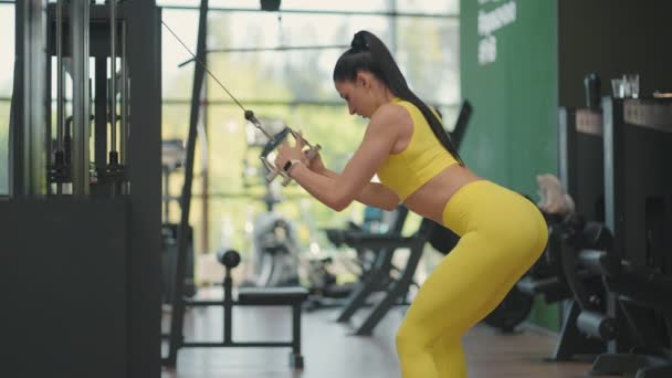 Sarı eşofmanlı genç bir İspanyol kadın, üst geçitte egzersiz yapıyor. Sırtını ve omuzlarını eğitmek için yukarıdan çelik bir ip çekiyor. Bir kadın spor salonunda sırtını ve omuzlarını çalıştırıyor. - Video, Çekim
