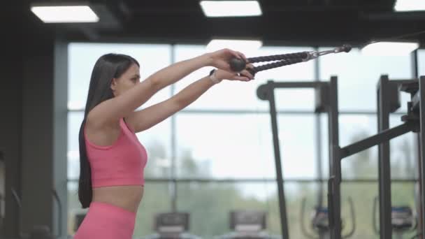 A brunette woman in a pink suit pulls a rope in a crossover trainer to her chest. Back workout in a trainer. Professional woman instructor - Footage, Video