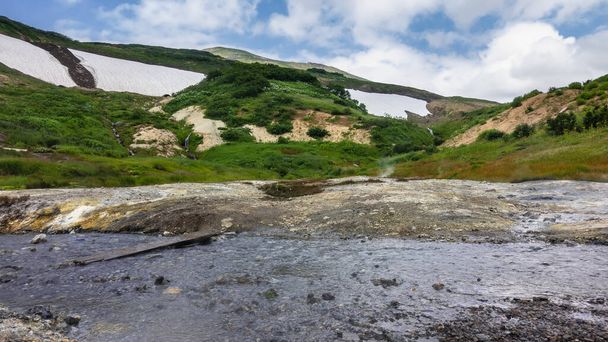Un ruisseau des sources chaudes coule le long d'un lit rocheux. Il y a des dépôts de soufre sur le rivage. Des brins de vapeur au-dessus du sol. Sur le flanc de la montagne, il y a de la végétation verte, de la neige fondue. Kamchatka - Photo, image