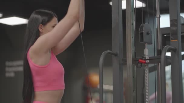 A hispanic brunette woman in a pink suit pulls a rope in a crossover with her hands to train her shoulders. Shoulder workout in a trainer. Professional woman instructor - Footage, Video