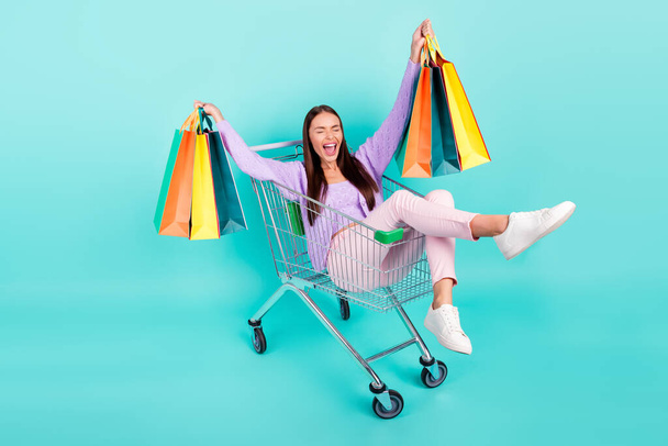 Photo of sweet funky woman dressed purple clothes holding bags riding shopping tray laughing smiling isolated teal color background - Foto, Imagem
