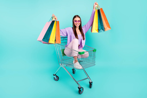 Fotografie turistická dáma jízda vozík držení tašky supermarket nosit sluneční sklo fialový svetr izolované teal barva pozadí - Fotografie, Obrázek